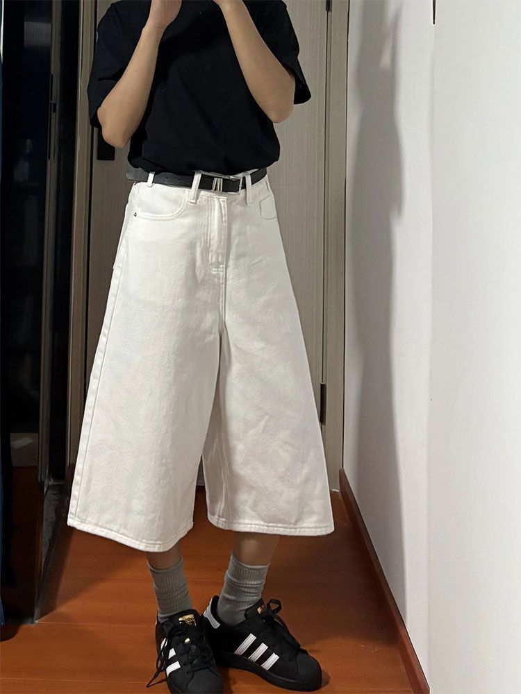 A person wearing a black short-sleeve t-shirt and high-waisted, wide-leg white denim culottes, styled with a black belt, gray socks, and black sneakers with white stripes.