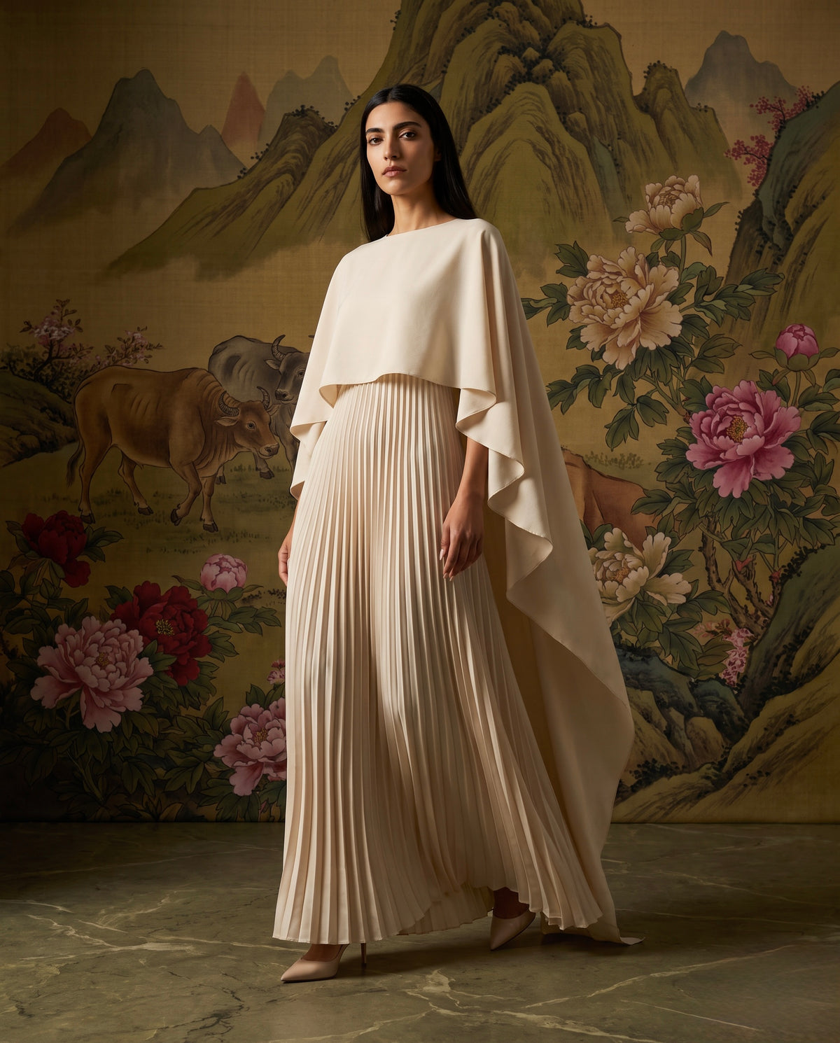 A woman models a floor-length cream pleated gown with a draped cape-like overlay and wide sleeves, standing in front of a large traditional-style landscape tapestry with mountains, flowers, and a deer.